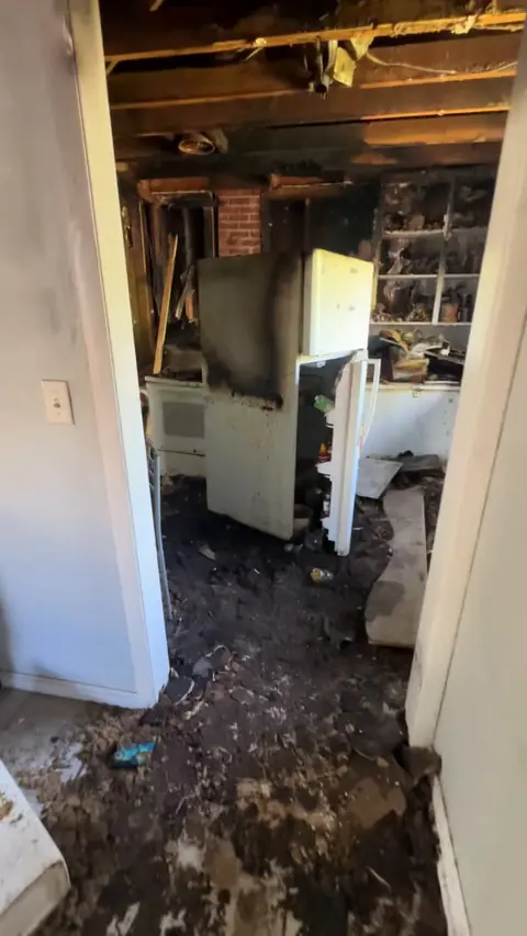 Kitchen seen through a doorway, with the ceiling open and burned debris on the floor